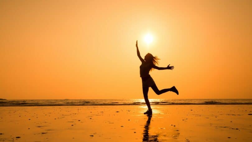 Imagem do pôr do sol em uma praia e em destaque a silhueta de uma moça agradecendo pela vida.