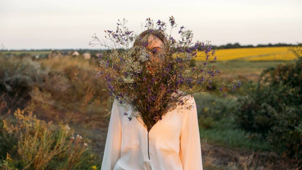 Imagem de um campo verde e em destaque uma mulher vestindo uma camisa branca celebrando a sua beleza interior e o amor próprio. Ela está feliz segurando um buquê de flores silvestres.