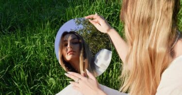 Imagem de uma moça de cabelos loiros e compridos olhando para um espelho. Ela está sentada em um gramado e busca refletir sobre o seu interior.
