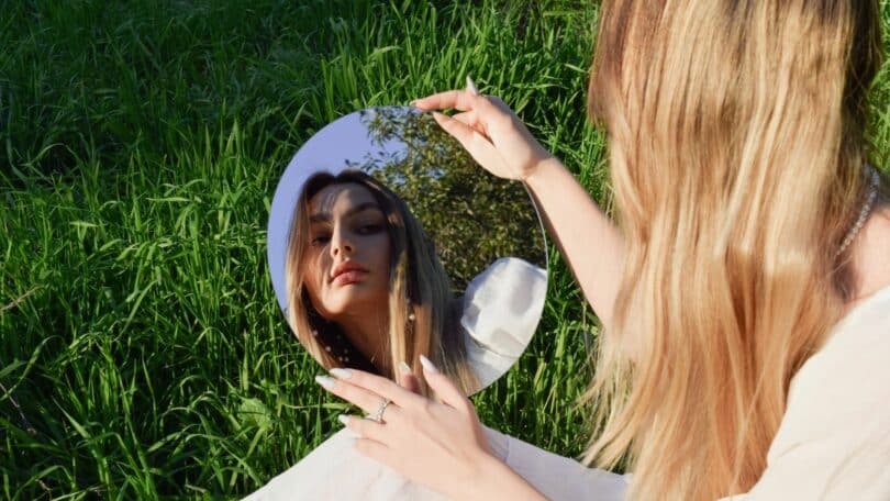 Imagem de uma moça de cabelos loiros e compridos olhando para um espelho. Ela está sentada em um gramado e busca refletir sobre o seu interior.