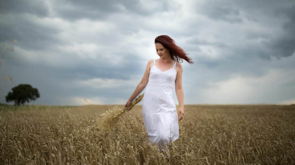 Imagem de um campo de trigo e em destaque uma mulher de cabelos ruivos, usando um vestido branco segurando um ramo da planta.
