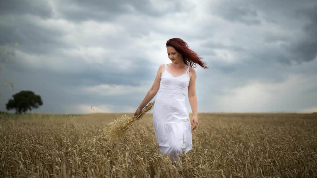 Imagem de um campo de trigo e em destaque uma mulher de cabelos ruivos, usando um vestido branco segurando um ramo da planta.