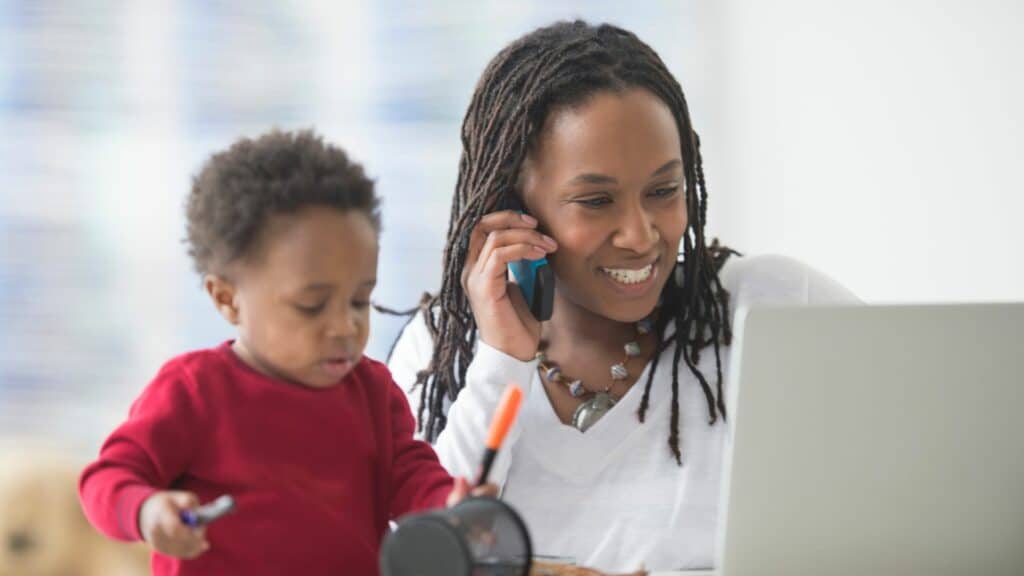 Imagem de uma mulher negra e seu filho. Ela está trabalhando com ele ao seu lado.
