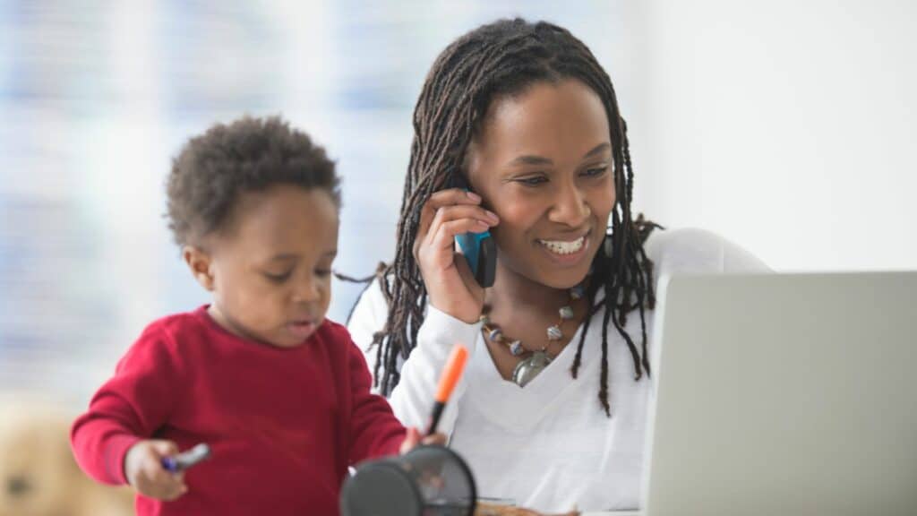 Imagem de uma mulher negra e seu filho. Ela está trabalhando com ele ao seu lado.
