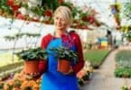 Imagem de uma floricultura e em destaque uma mulher mãe e empreendedora segurando dois casos de plantas.