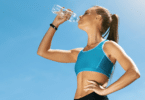 Mulher tomando garrafinha d'água após correr. Ela está vestindo roupas esportivas, em fundo azul.