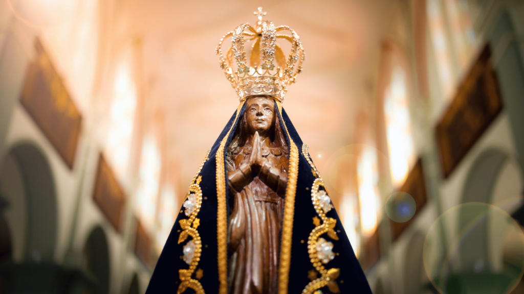 Imagem de Nossa Senhora Aparecida. No seu fundo, há uma igreja iluminada pela luz do sol.