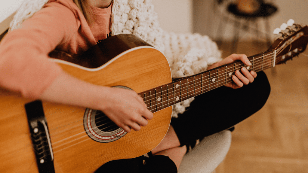 Mulher tocando violão, enquanto está sentada de pernas cruzadas. 