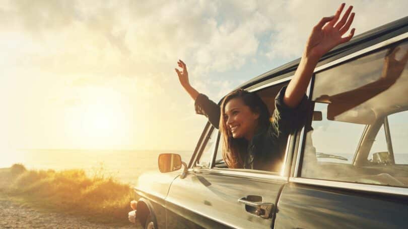 Imagem de um pôr do sol em uma praia. Em destaque um carro e uma mulher toda feliz, abrindo os braços na janela do carro mostrando que está vivendo a vida intensamente.