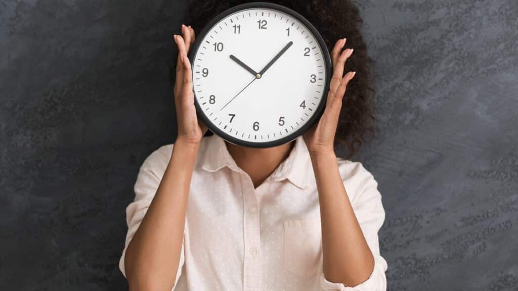 Imagem de fundo preto e em destaque uma mulher de cabelos ondulados segurando em frente ao seu rosto um relógio redondo, aplicando o conceito de gerenciamento de tempo.
