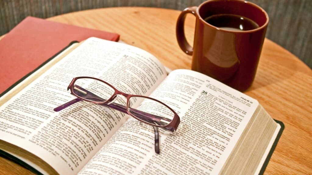 Imagem de uma bíblia sobre uma mesa, uma xícara de café e um óculos de grau. Uma pessoa está fazendo a sua devoção matinal.