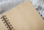 Caderno sobre toalha branca com renda em fundo de madeira.