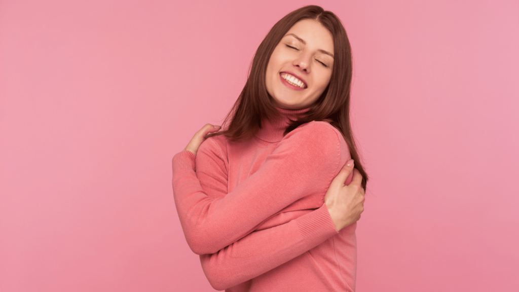 Mulher se abraçando em fundo rosa. 