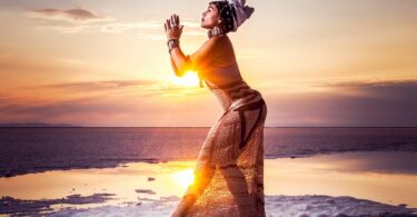 Mulher com as mãos juntas, em posição de Yoga, em frente do por do sol.