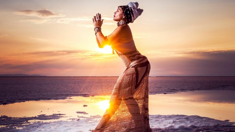 Mulher com as mãos juntas, em posição de Yoga, em frente do por do sol.