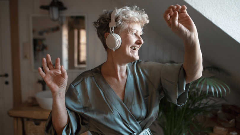 Mulher sênior feliz com fones de ouvido dançando em casa, relaxando e conceito de autocuidado.