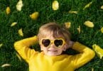 Imagem de uma criança loira, usando uma camiseta de manga longa e um óculos em formato de coração na cor amarela. Ela está deitada em um gramado coberto com folhas amarelas caídas de uma árvore.