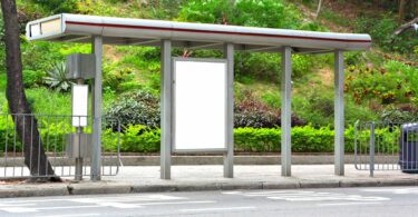 Imagem de um ponto de ônibus vazio. Ao fundo dele, imagens de um jardim.