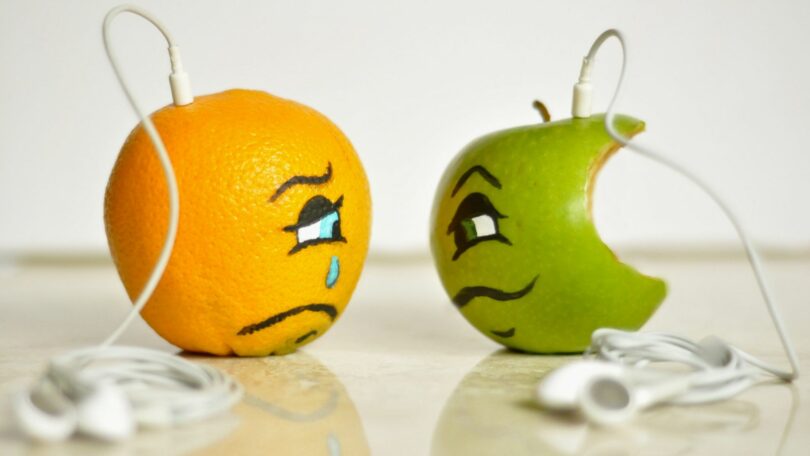Imagem de uma laranha e uma maçã verde. Uma está bonita e a outra já está mordida. Ambas usam um fone de ouvido e estão representando o sentimento de inveja.