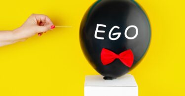 Imagem de fundo amarelo e em destaque um balão preto com o texto Ego escrito sobre ele e mão de uma mulher que vai estourar o balão com agulha. A imagem traz os conceitos de orgulho e egoísmo.