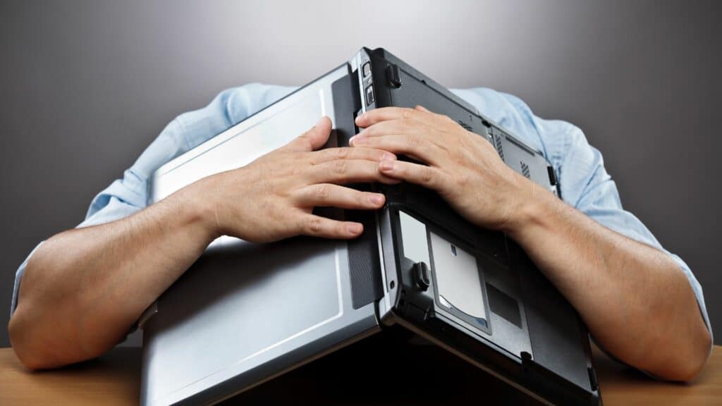 Imagem de fundo cinza e em destaque um homem sentado, colocando a sua cabeça dentro do seu notebook, representando uma exaustão profissional.