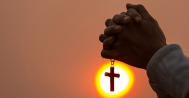 Mãos juntas, segurando uma corrente com cruz, em sinal de oração. O fundo está iluminado de amarelo e laranja.