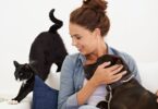 Imagem de fundo branco e em destaque uma moça de cabelo preso, sentanda brincando com um gato e um cachorro, seus animais de estimação, mostrando a prática da convivência.