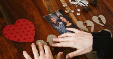 Imagem de uma mesa de madeira e sobre ela o retrato de um casal, um coração vermelhor inteiro e outros dois corações de papelão cortado ao meio. Uma mão tenta juntar esse coração. Conceito de separação de casal, infidelidade.
