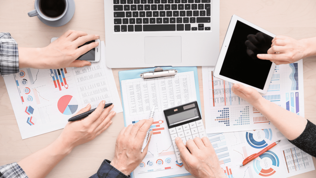 Pessoas trabalhando juntas em uma mesa, com gráficos expostos, notebooks, tablets e celulares. 