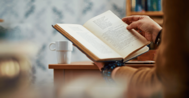Pessoa segurando livro enquanto o lê, próximo de mesa com xícara de café.