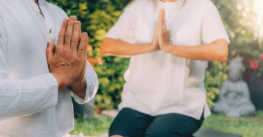 Duas pessoas sentadas em tapete sobre o ar livre, um jardim. Elas estão em posição de meditação, com as mãos juntas.