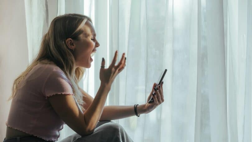 Mulher loira de cabelos lisos está com expressão de raiva e olha para o celular. Ela está sentada e posicionada de lado na imagem e, ao fundo, há uma cortina branca. Ela usa uma blusa rosa e veste calça jeans.