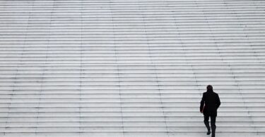 Um homem de roupa preta e com uma pasta vermelha na mão sobe sozinho uma grande escadaria branca. Ele está posicionado no cantor inferior esquerdo da imagem. A maior parte da foto é composta pela escadaria que, em contraste com o homem subindo e ocupando um pequeno espaço da imagem, dá a sensação de solidão e vazio.