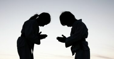 As silhuetas de duas crianças compostas por uma menina e um menino estão de frente uma para a outra e elas se curvam em sinal de reverência e respeito. Ao fundo e atrás das silhuetas, há um céu azul claro.