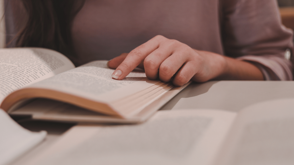 Uma mulher lendo um livro. A imagem foca na mão esquerda de uma mulher, apontando o dedo indicador em um página e acompanhando a leitura.