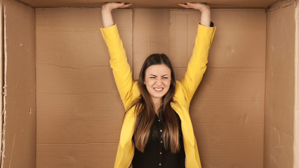 Mulher de camisa preta e blazer amarelo está sentada dentro de uma caixa de papelão. Ela levanta os dois braços, toca a parte de cima da caixa, representando "alcançar o limite". Ela tem uma expressão de insatisfação.