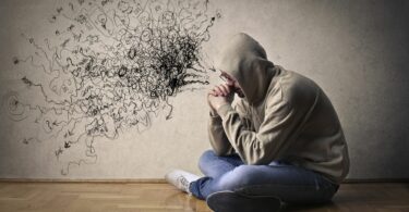 Imagem de um homem sentado, cabisbaixo pensando em seus sentimentos e desejos. Ao fundo uma parede bege com alguns rabiscos, representando os seus pensamentos.