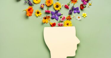 Imagem de fundo verde e em destaque a silhueta de uma cabeça humana e dentro dela, saem algumas flores coloridas, trazendo o conceito de saúde mental.
