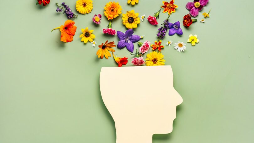 Imagem de fundo verde e em destaque a silhueta de uma cabeça humana e dentro dela, saem algumas flores coloridas, trazendo o conceito de saúde mental.