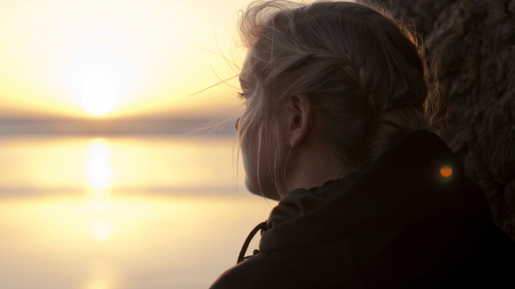 Imagem de uma mulher triste e com semblante apagado, olhando para o pôr do Sol em frente a um lago.
