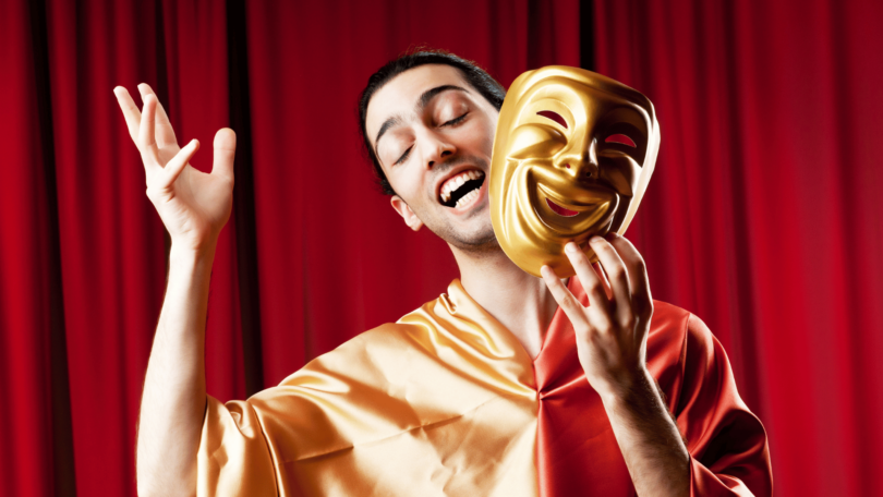 Imagem de um ator masculino em cima do palco de um teatro. Ele está de olhos fechados, sorrindo e segurando em uma das mãos uma máscara dourada.