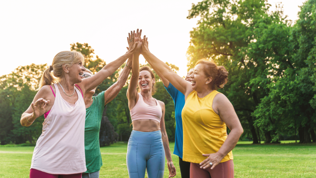 Um grupo de cinco mulheres que fazem exercícios estão rindo e juntando as mãos no alto, indicando uma união. Elas vestem roupas esportivas leves em um parque. No fundo, há grama, árvores e a luz do sol desfocados.