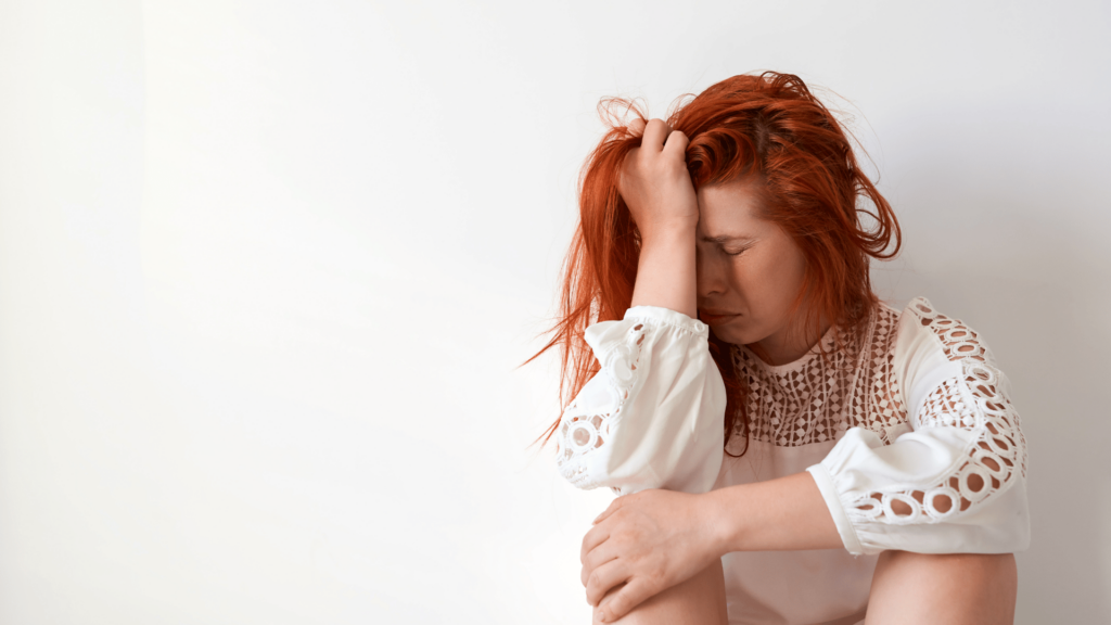 Mulher ruiva está sentada no chão com uma mão na cabeça e outra apoiada nos joelhos. Ela expressa uma emoção negativa, como estresse ou tristeza. Ela veste uma blusa branca e há um fundo branco atrás.