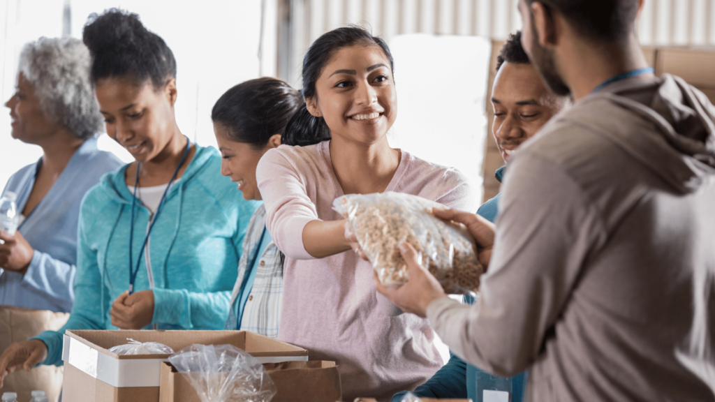 Uma mulher jovem de cabelos castanhos está no foco da imagem e exerce uma atividade voluntária. Ela entrega um alimento a outro voluntário e sorri. Há outros cinco voluntários interagindo ao seu lado.
