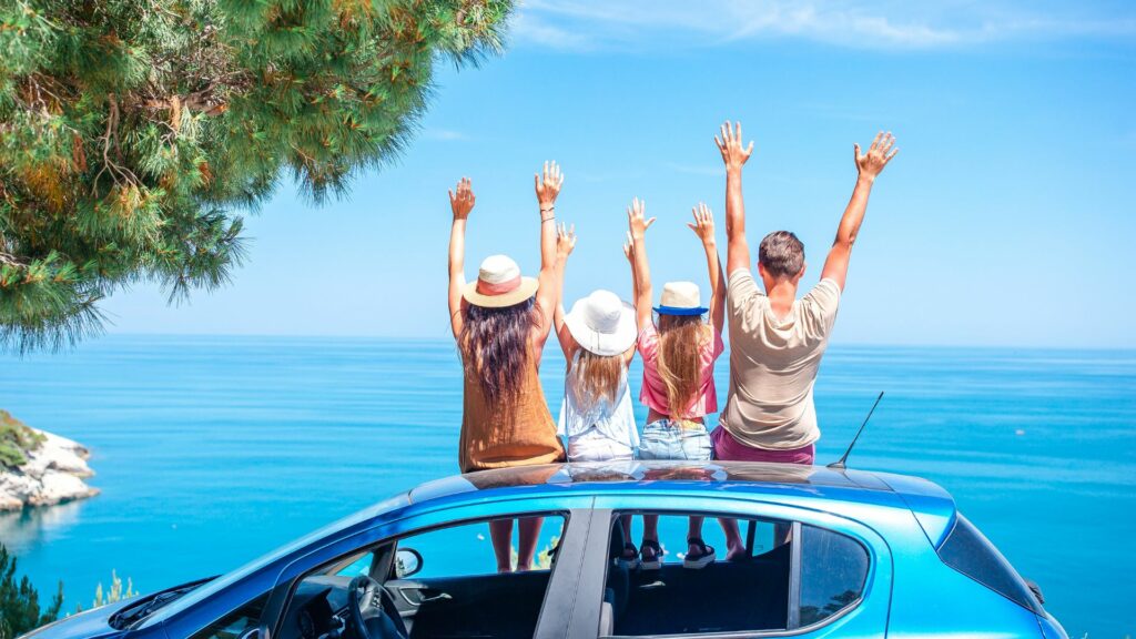 Imagem de uma família, pais e filhas fazendo uma viagem. Eles estão sentadores sobre o carro, de braços levantandos comtemplado a imensidão do mar.
