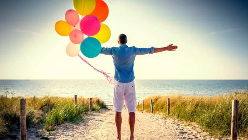 Imagem de fundo de um lindo mar azul e em destaque um homem usando uma bermuda branca e um camisa jeans. Ele está contemplando a beleza do mar e a felicidade da vida. Ele ainda está segurando em uma das mãos, vários balões coloridos.