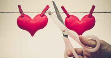 Imagem de um varal feito de barbante. Nele estão pendurados dois corações na cor vermelho, feitos de feltro. Uma mão masculina, segurando uma tesoura, corta este varal, simbolizando o fim do amor.
