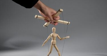 Imagem do braço de um homem puxando os cordões de uma marionete em um fundo cinza, simbolizando o conceito de manipulação social, falta de liberdade.