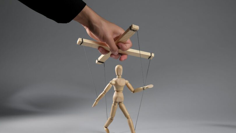 Imagem do braço de um homem puxando os cordões de uma marionete em um fundo cinza, simbolizando o conceito de manipulação social, falta de liberdade.