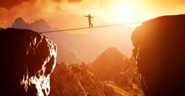 Imagem de um homem andando e se equilibrando em uma corda que liga uma pedra a outra, simbolizando o conceito de desafio e oportunidade.